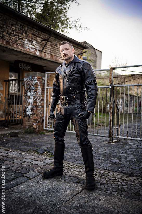 SM Master Andre alias DominusBerlin in Voll Leder Uniform mit Jacke Hose und Stiefeln in Industrie Umgebung mit Eisen Tor und Halle mit Graffiti besprueht