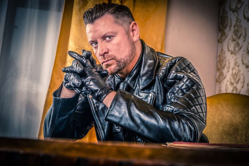 The dominus, upper-body photograph, staring directly at the camera, clad in black leather jacket and folding his hands with black leather gloves.