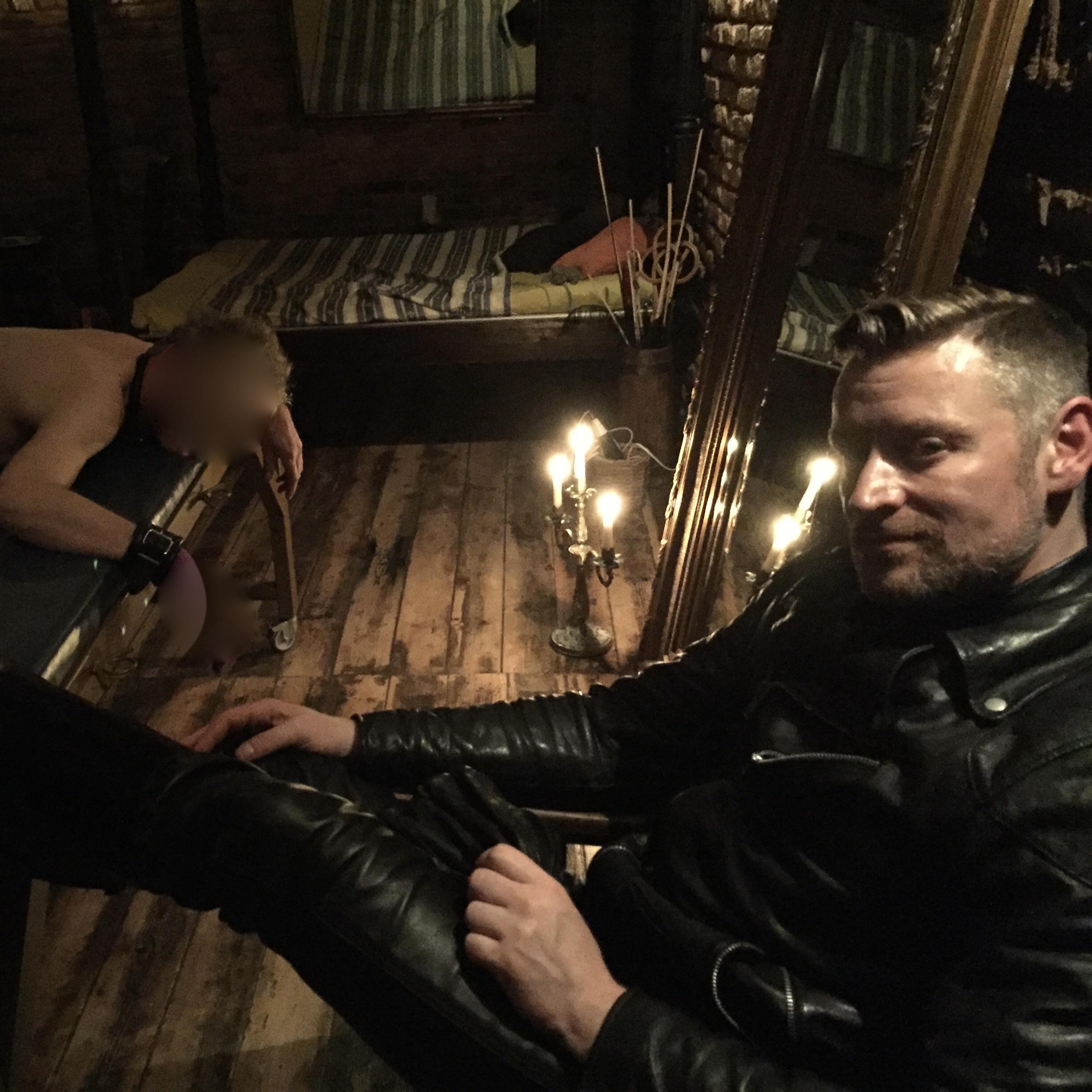 SM Master Dominus Andre sitzt in Leder auf seinem Stuhl und sieht den gefesselten nackten Sklaven an