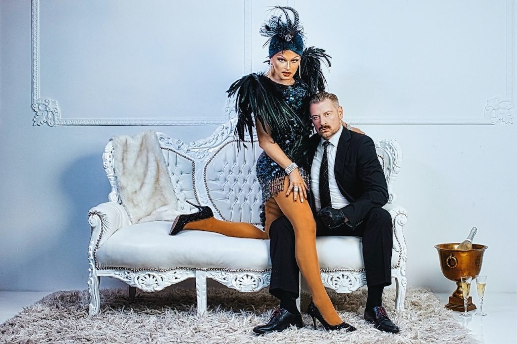 Fetisch Modell Alex Varlan auf Schoss von SM Master DominusBerlin alias Master Andre in gehobenem Ambiente auf Sofa sitzend mit Anzug und Diven Outfit