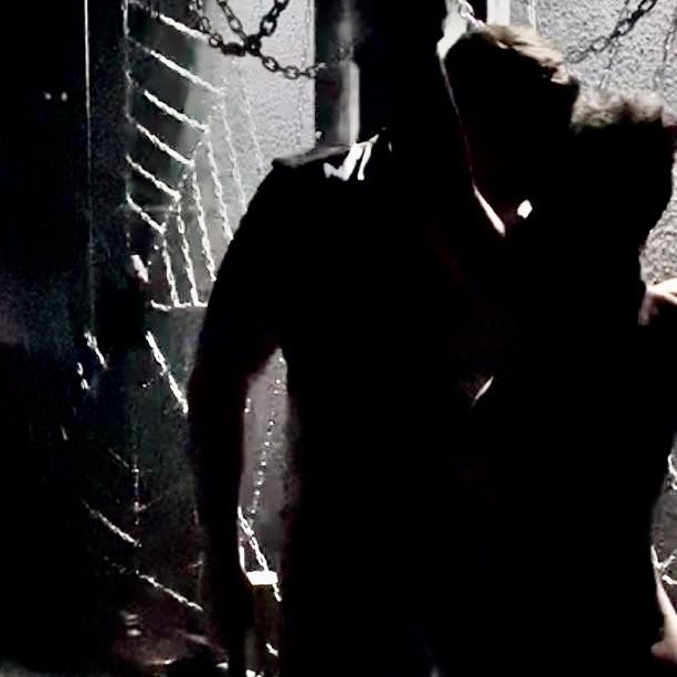 Die in Ketten gelegte Sklavin steht devot vor dem Dominus. Im Hintergrund ist das Ketten Spinnennetz im SM Studio zu sehen.