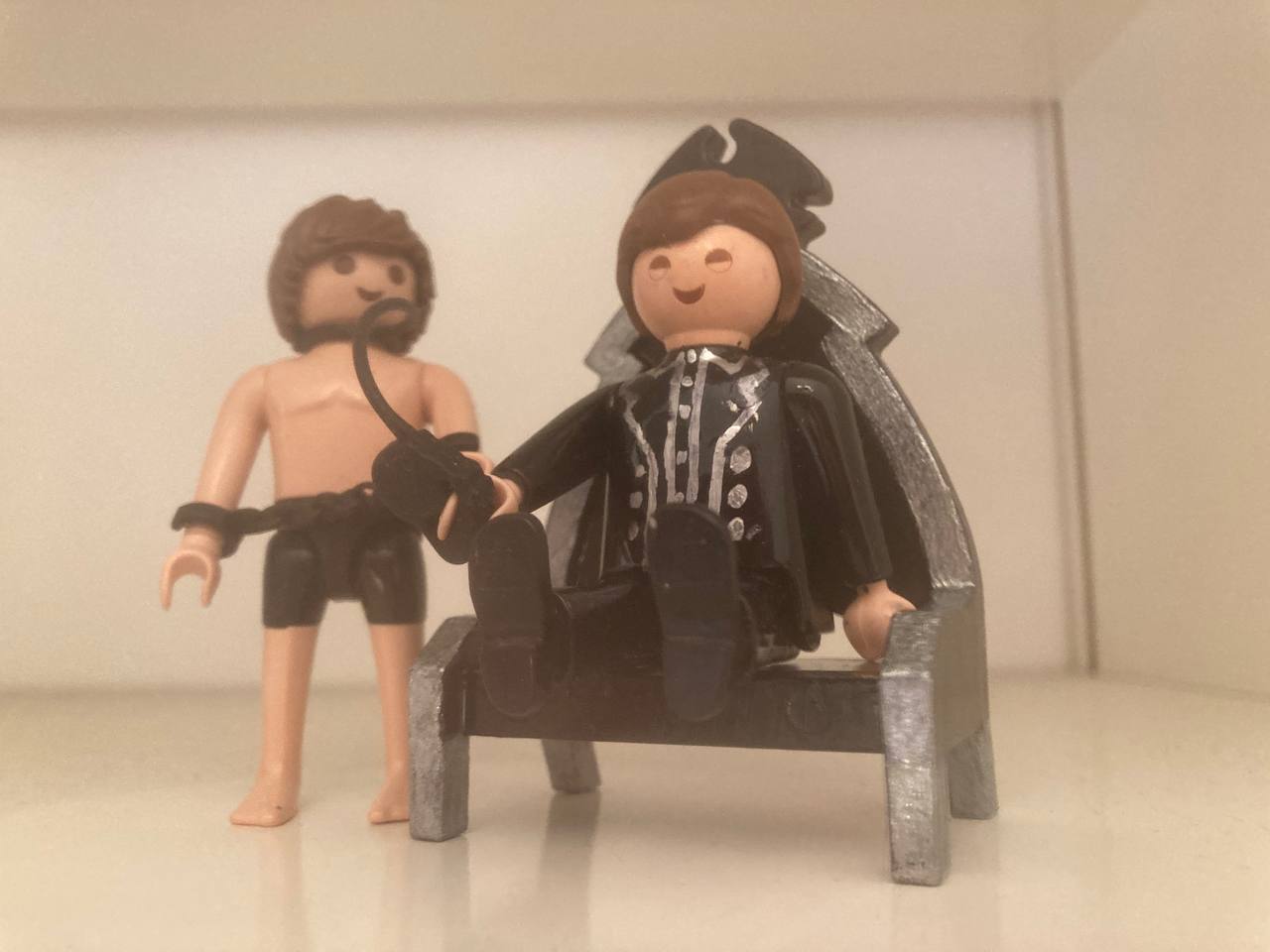 Zwei Playmobil Figuren stellvertretend für SM Master Andre alias Der Dominus in schwarzer Kleidung auf seine, Thron und einem nackten, gefesselten Sklaven, der auf die Peitsche wartet